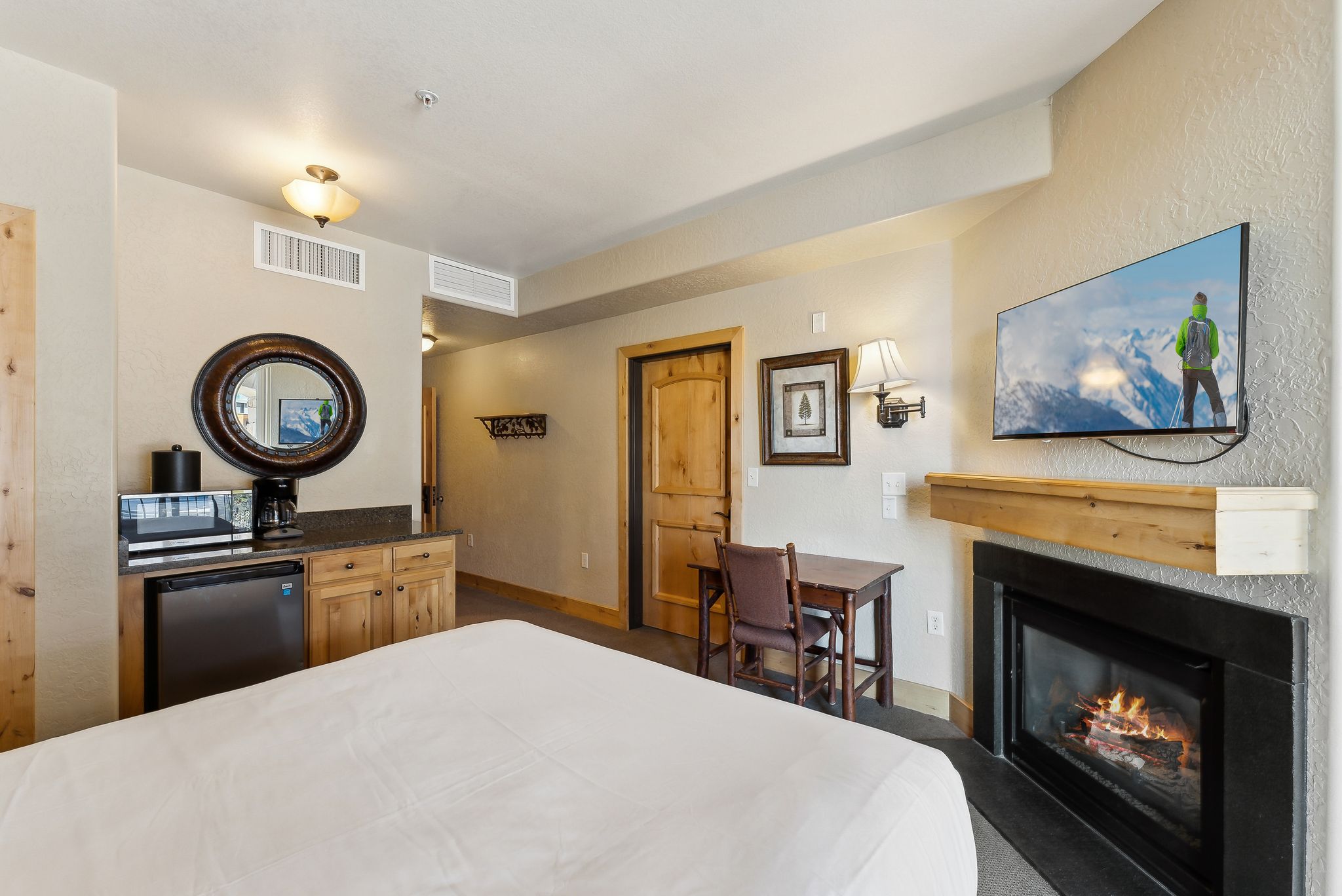 Room featuring a built-in fireplace with a natural wood mantel, a wall-mounted flat-screen television, a compact kitchenette with a built-in refrigerator and microwave, and a wooden desk with a chair