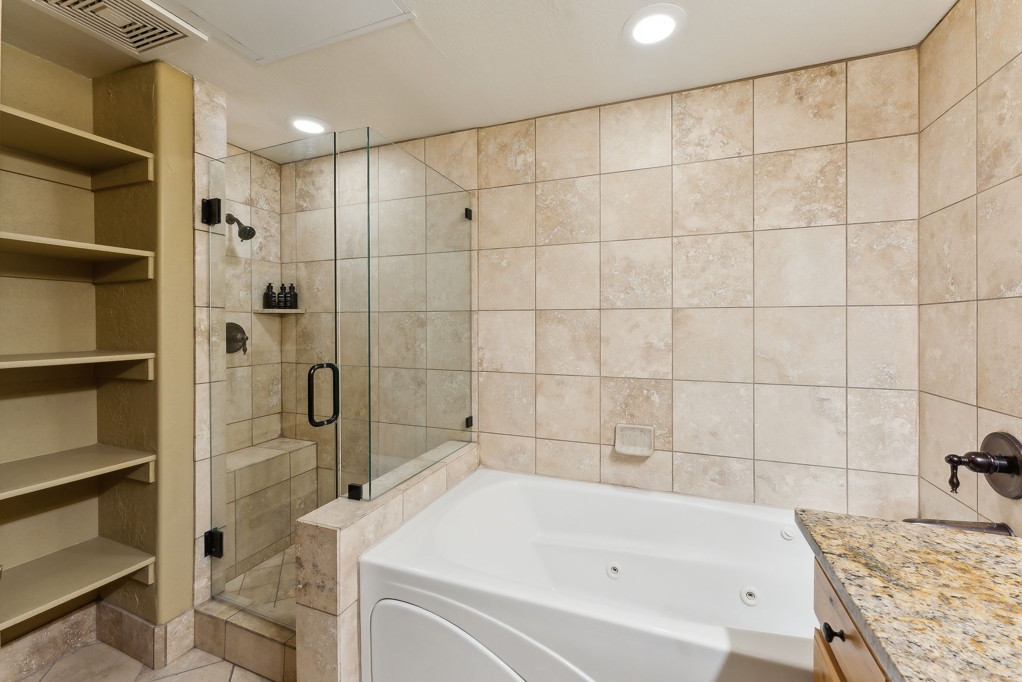 Tiled bathroom featuring a glass-enclosed shower with a built-in bench, a jetted soaking tub, built-in shelving, and a granite-topped vanity
