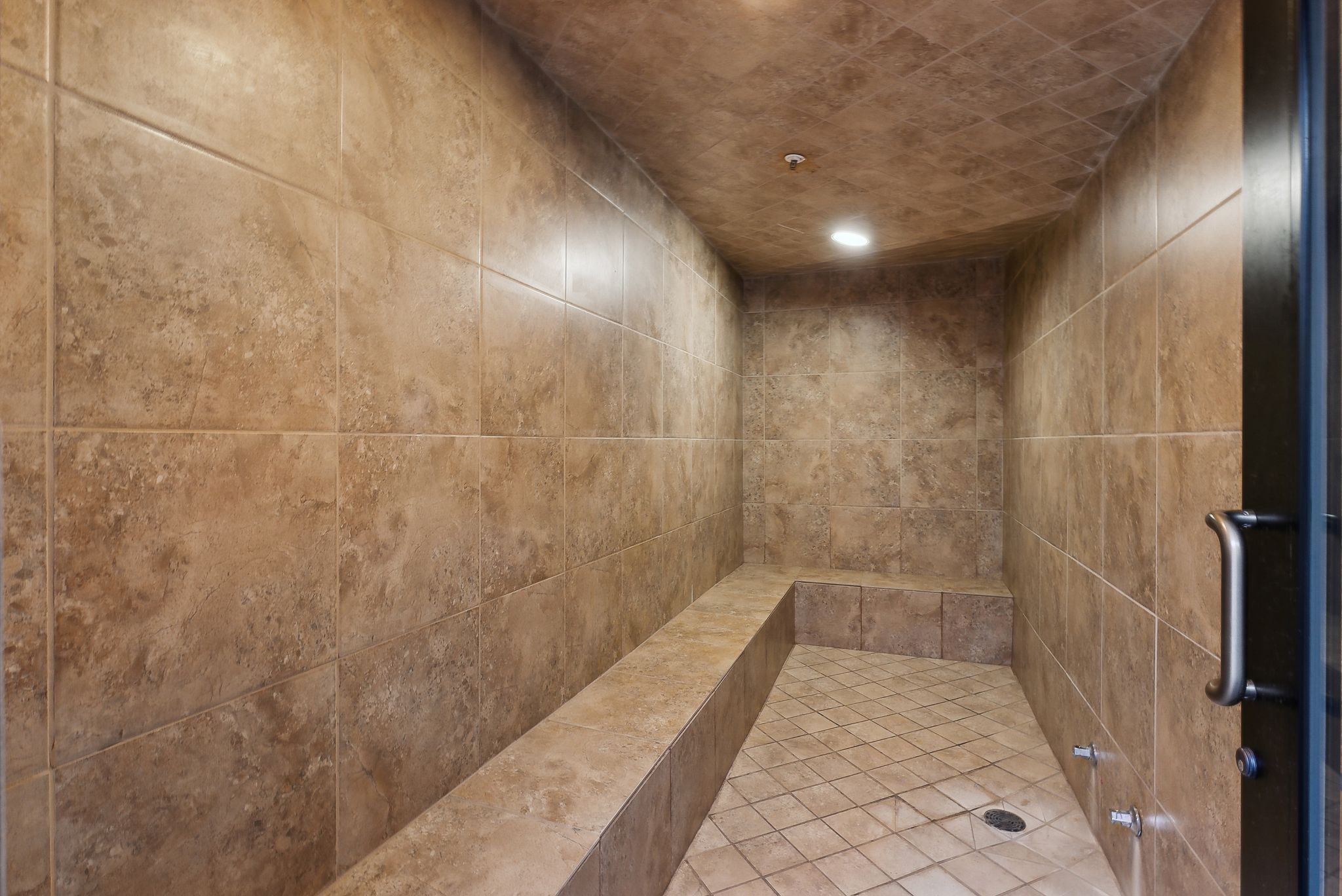 Tiled steam room featuring a built-in bench, recessed lighting, floor drain, and dark-framed glass door with a metal pull handle
