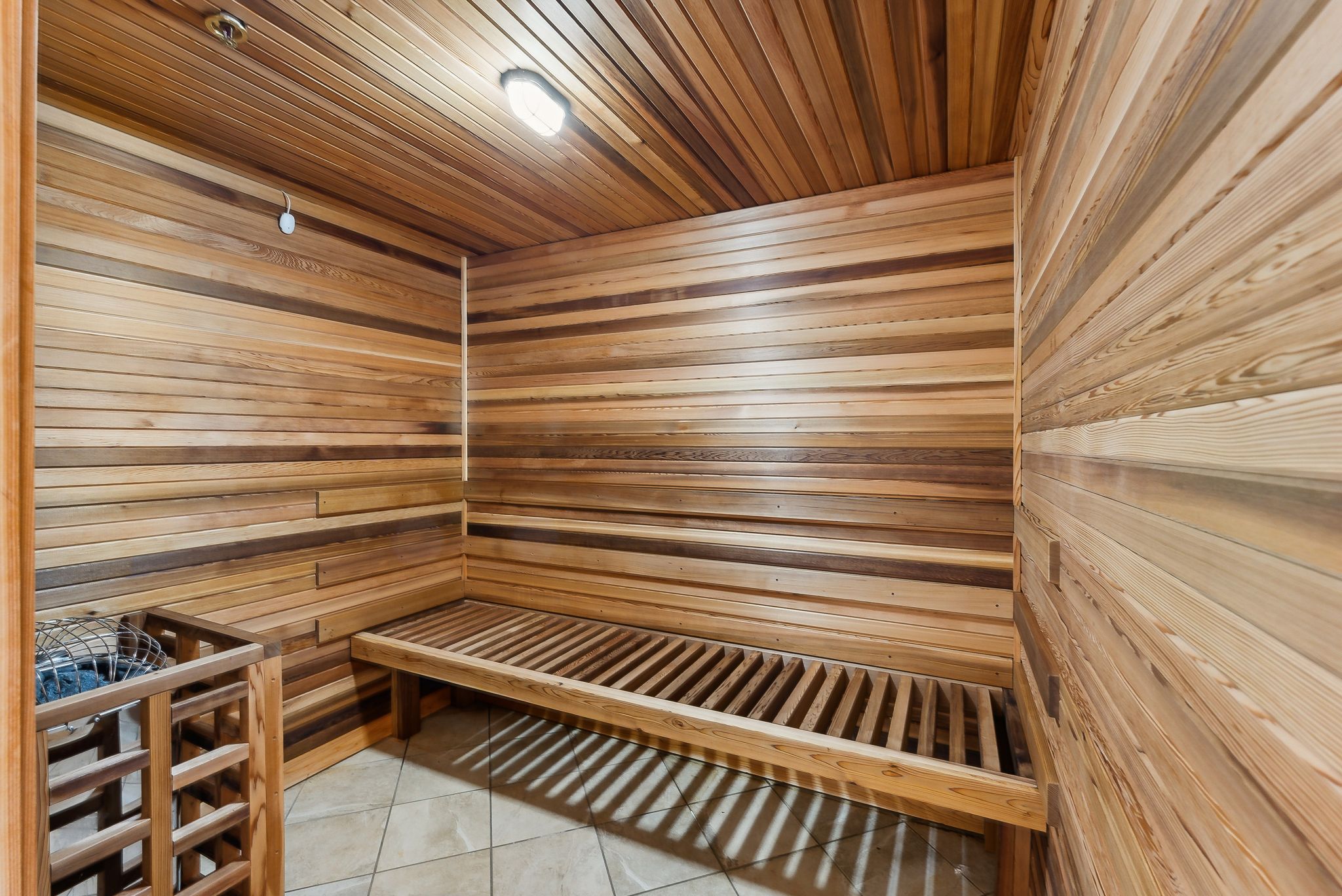 Sauna interior featuring horizontal wood paneling, slatted wood bench seating, tile flooring, and a built-in heater with a protective wood surround