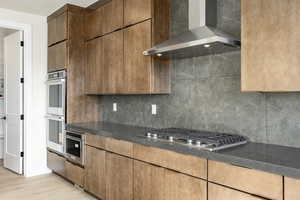 Kitchen with wall chimney range hood, backsplash, stainless steel appliances, light wood-type flooring, and modern cabinets