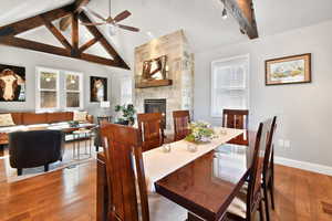 Dining area with ceiling fan, a stone fireplace, wood finished floors, vaulted ceiling with beams, and baseboards