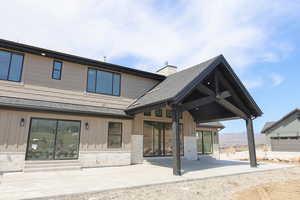 Rear view of house featuring a patio, board and batten siding, a shingled roof, and stone siding