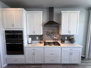 Kitchen featuring dobule oven black, wall chimney range hood, stainless steel gas stovetop, white cabinets, and decorative backsplash