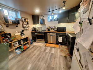 Kitchen with wall chimney exhaust hood, dark cabinets, tasteful backsplash, light wood-type flooring, and stainless steel appliances