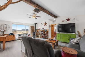 Living area with a wood stove, lofted ceiling with beams, a ceiling fan, visible vents, and wood finished floors