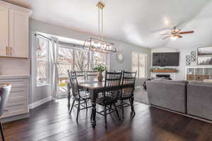 Dining room with baseboards, vaulted ceiling, a fireplace, dark wood-style floors, and ceiling fan with notable chandelier