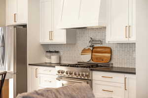 Kitchen with white cabinets, appliances with stainless steel finishes, dark countertops, custom range hood, and backsplash