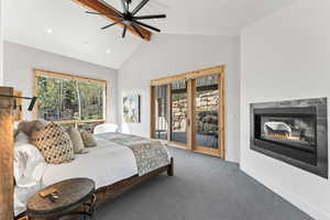 Carpeted bedroom featuring access to exterior, vaulted ceiling with beams, baseboards, a glass covered fireplace, and a ceiling fan