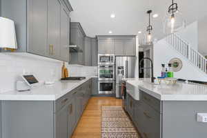 Kitchen featuring an island with sink, light stone counters, stainless steel appliances, decorative backsplash, and gray cabinetry