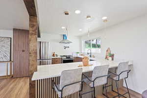 Kitchen with wall chimney exhaust hood, a peninsula, a kitchen bar, light wood-style floors, and appliances with stainless steel finishes
