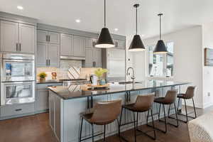 Kitchen featuring gray cabinets, tasteful backsplash, dark wood-style flooring, double oven, and a kitchen island with sink