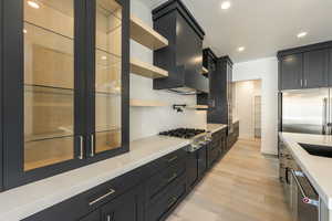 Kitchen featuring appliances with stainless steel finishes, wall chimney exhaust hood, light wood-style flooring, dark cabinetry, and open shelves