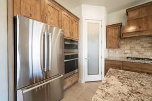 Kitchen featuring light stone counters, stainless steel appliances, premium range hood, decorative backsplash, and brown cabinetry
