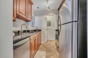 Kitchen featuring appliances with stainless steel finishes, a sink, brown cabinetry, light tile patterned floors, and dark stone countertops