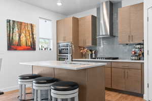 Kitchen featuring tasteful backsplash, a kitchen breakfast bar, wall chimney range hood, light countertops, and a sink