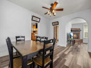 Dining space featuring a ceiling fan, light wood-type flooring, arched walkways, baseboards, and a brick fireplace