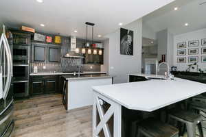Kitchen with a spacious island, wall chimney range hood, stainless steel microwave, tasteful backsplash, and light wood-type flooring
