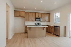 Kitchen featuring light wood finished floors, a center island, brown cabinetry, light countertops, and appliances with stainless steel finishes