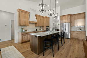 Kitchen with an island with sink, premium range hood, light wood-style floors, appliances with stainless steel finishes, and a breakfast bar area