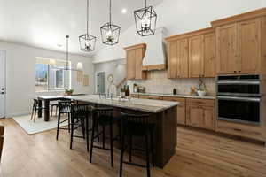 Kitchen featuring an island with sink, stainless steel double oven, decorative backsplash, a chandelier, and light wood-style flooring