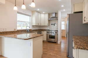 Kitchen with wall chimney range hood, a sink, light wood-style floors, a peninsula, and appliances with stainless steel finishes