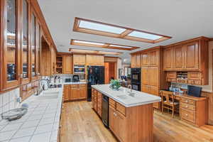 Kitchen featuring open shelves, a sink, brown cabinetry, black appliances, and a center island with sink