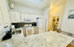 Kitchen with white appliances, a sink, a peninsula, light stone counters, and white cabinetry