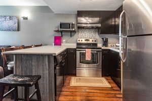 Kitchen with wall chimney exhaust hood, stainless steel appliances, a peninsula, a kitchen breakfast bar, and light countertops