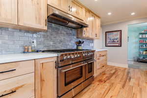 Kitchen with under cabinet range hood, light brown cabinetry, double oven range, baseboards, and ornamental molding