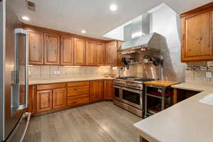 Kitchen with wall chimney exhaust hood, light wood finished floors, tasteful backsplash, brown cabinets, and appliances with stainless steel finishes