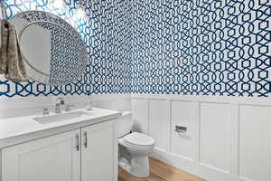 Bathroom with toilet, wainscoting, wallpapered walls, wood finished floors, and vanity