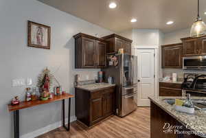 Kitchen with light stone counters, stainless steel appliances, dark brown cabinets, light wood-type flooring, and pendant lighting