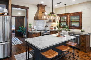 Kitchen with light stone counters, dark wood-type flooring, a kitchen bar, appliances with stainless steel finishes, and custom exhaust hood