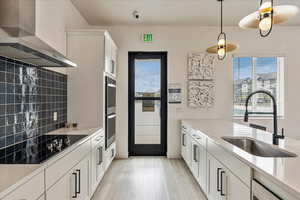 Kitchen with wall chimney exhaust hood, black electric cooktop, light wood-style flooring, a sink, and backsplash