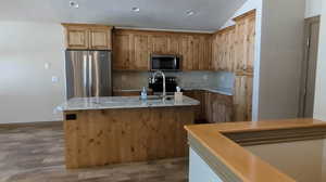 Kitchen with stainless steel appliances, a kitchen island with sink, vaulted ceiling, wood finished floors, and light stone counters