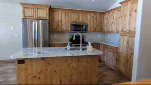 Kitchen with appliances with stainless steel finishes, wood finished floors, a sink, a center island with sink, and vaulted ceiling