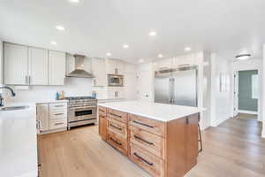 Kitchen with light wood-style flooring, a sink, built in appliances, wall chimney range hood, and a kitchen island