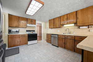 Kitchen with brown cabinetry, appliances with stainless steel finishes, a sink, light countertops, and under cabinet range hood