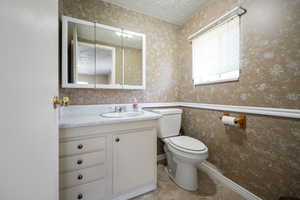 Half bathroom with vanity, toilet, wallpapered walls, and a textured ceiling