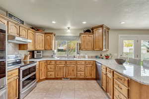 Kitchen featuring light stone counters, a sink, appliances with stainless steel finishes, a peninsula, and under cabinet range hood