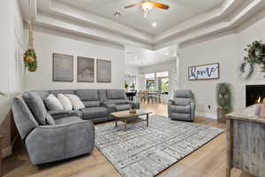 Living area with recessed lighting, a tray ceiling, a warm lit fireplace, a ceiling fan, and light wood-style floors