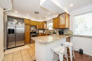Kitchen with stainless steel appliances, a peninsula, backsplash, brown cabinets, and light stone countertops