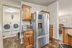 Kitchen with washing machine and clothes dryer, dark countertops, brown cabinets, stainless steel fridge with ice dispenser, and dark wood-style flooring
