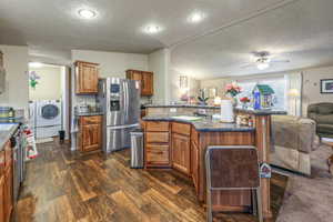 Kitchen with open floor plan, independent washer and dryer, stainless steel fridge with ice dispenser, brown cabinetry, and dark wood-type flooring