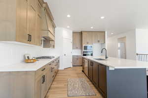 Kitchen with stainless steel appliances, a sink, light wood finished floors, tasteful backsplash, and a center island with sink