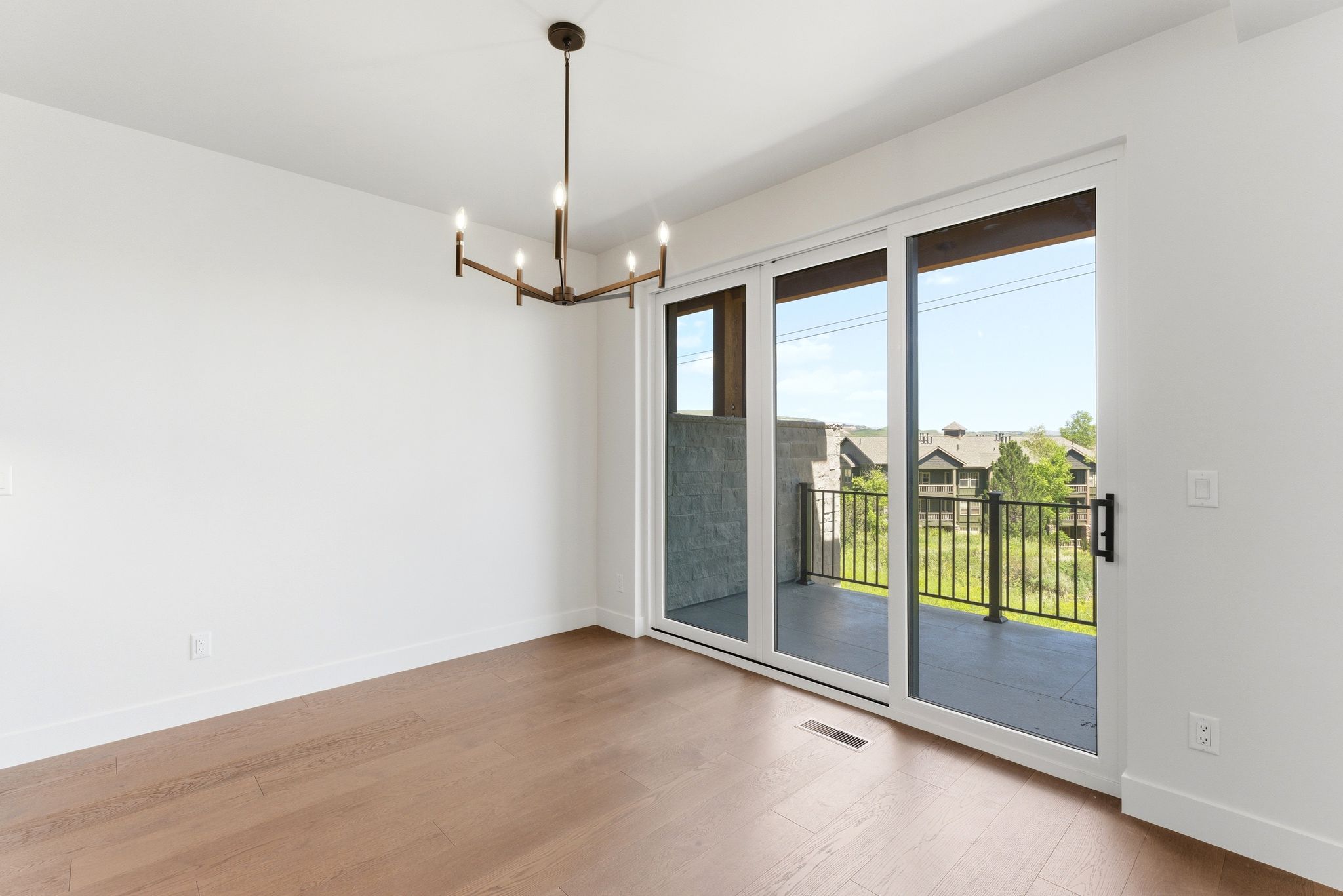 Spacious room featuring wood-finish flooring, a modern chandelier, and large sliding glass doors opening to a private exterior balcony with metal railings and a stone-finish privacy wall