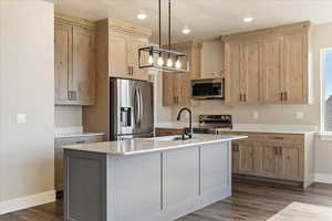 Kitchen featuring stainless steel appliances, light brown cabinetry, baseboards, a center island with sink, and dark wood-style floors