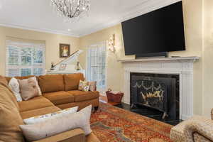 Living room with a premium fireplace, wood finished floors, a notable chandelier, and ornamental molding. Grand Piano and additional table seated area with view  windows on both ends of room.