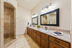 Full bath featuring stone tile floors, a sink, double vanity, and tiled shower